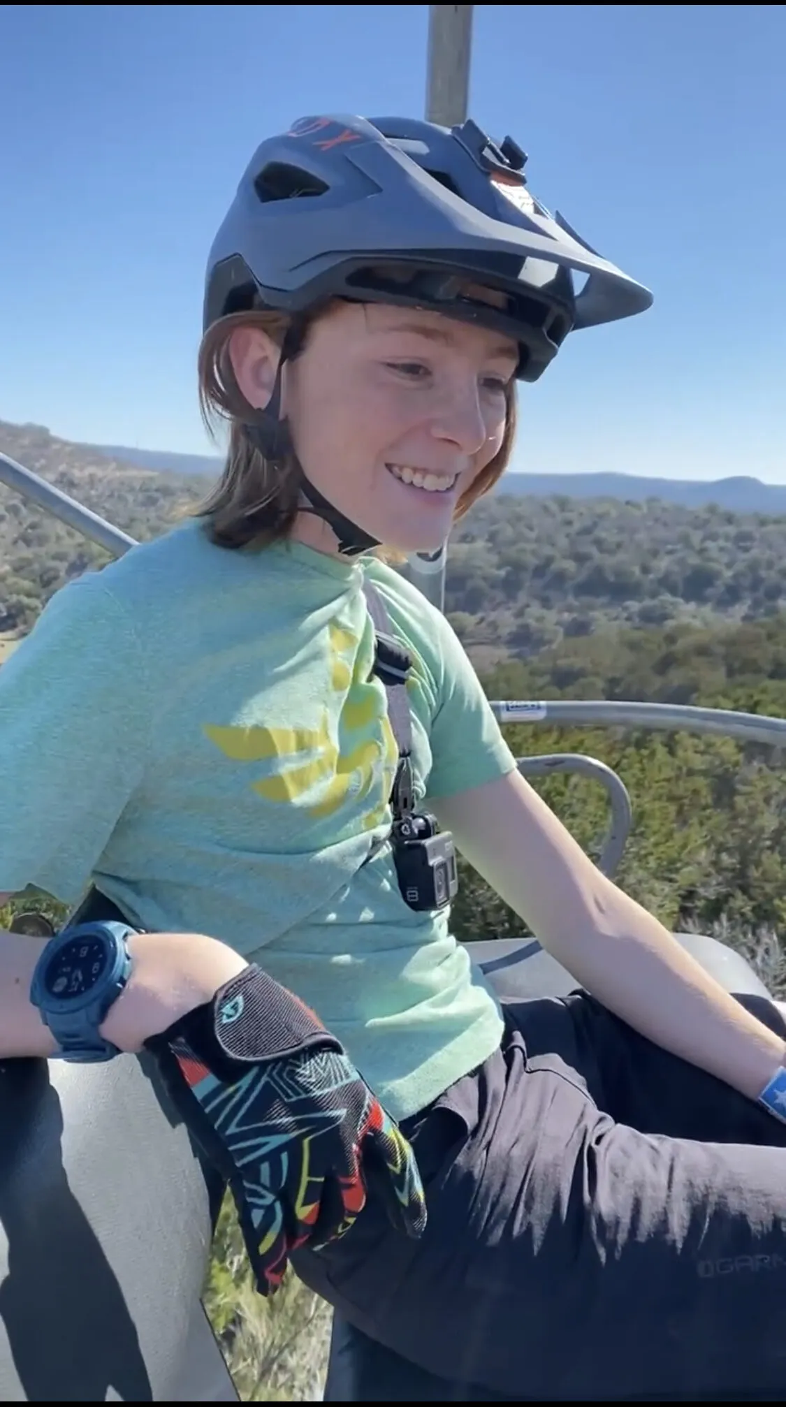 Jonas smiling on a chairlift in a mountain-bike helmet and riding gloves, with hills behind him